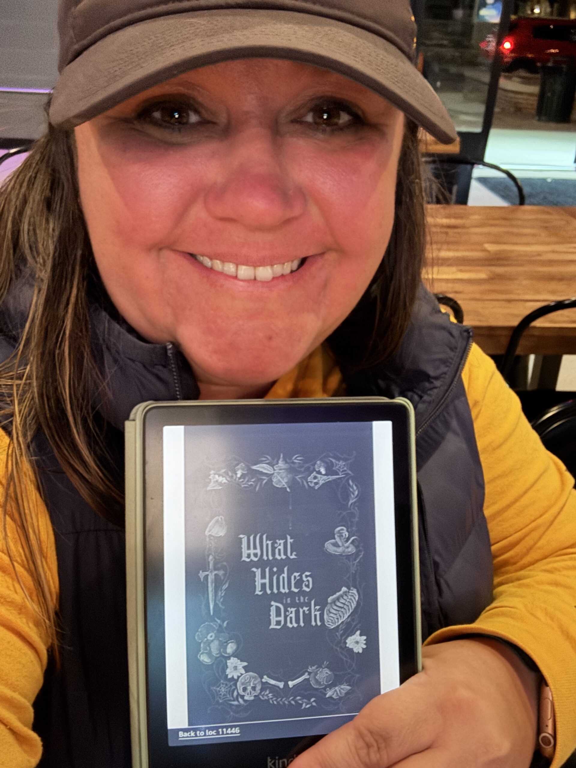 Image of author Laramie Cummings in a hat and showing a kindle that has the anthology, What we do in the dark